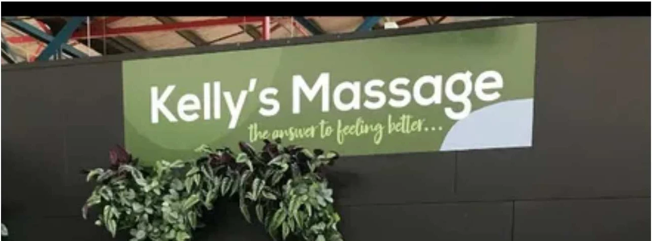Kelly's Massage green banner sign — 'Kelly's Massage / the answer to feeling better…' on a charcoal wall, exposed red steel truss above, trailing plants below