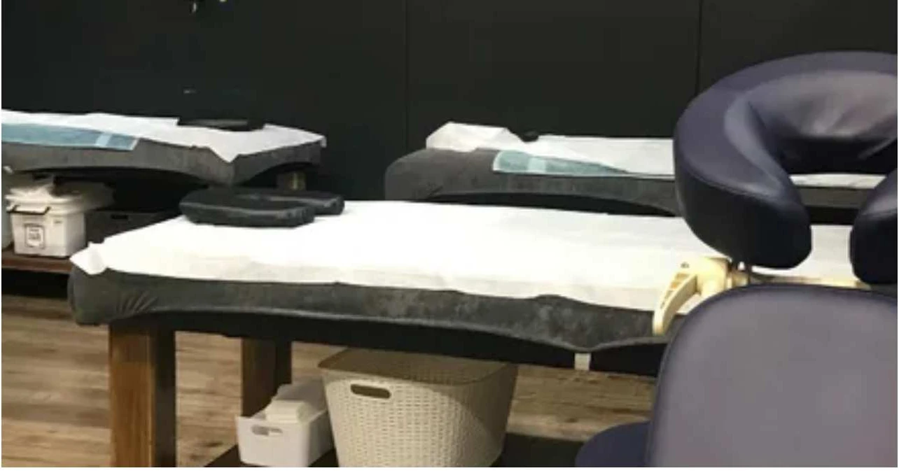 The Harvest Hall treatment room at Kelly's Massage — white-draped massage tables receding into a charcoal-walled room, a navy face-cradle chair in the foreground, honey timber floor