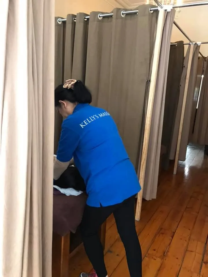 A Kelly's Massage therapist at work in a curtained treatment bay, royal-blue polo, a row of taupe bays behind
